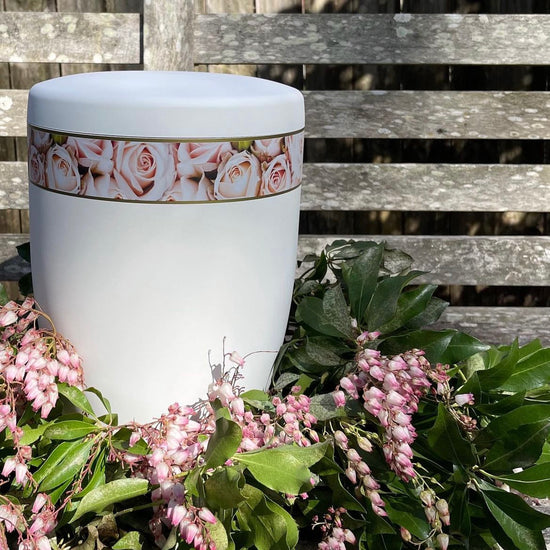 Beautiful white urn for ashes with its band of delicate pink roses set in a beautiful and simple wreath of delicate flowers and foliage.