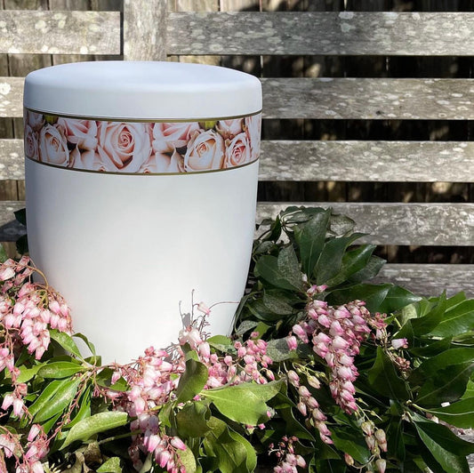Beautiful white urn for ashes with its band of delicate pink roses set in a beautiful and simple wreath of delicate flowers and foliage.