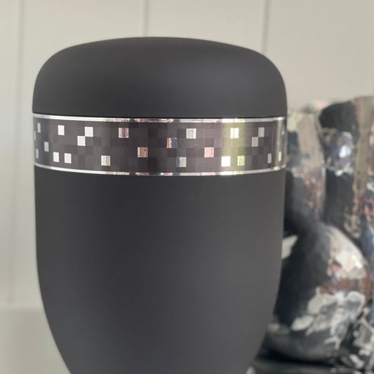 Stunning  urn for ashes adorned with a band of reflecting silver squares. This urn speaks of character strength and integrity. The urn is sitting on a white shelf with a silver vase in the background.