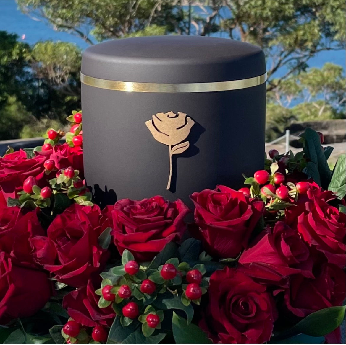 Elegant black funeral urn with a symbol of a golden rose set in a beautiful wreath of red roses.