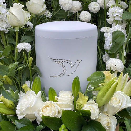 Elegant white urn for ashes adorned with a beautiful dove set in sparkling Swarovski crystals  in a bouquet of white lilies and roses.