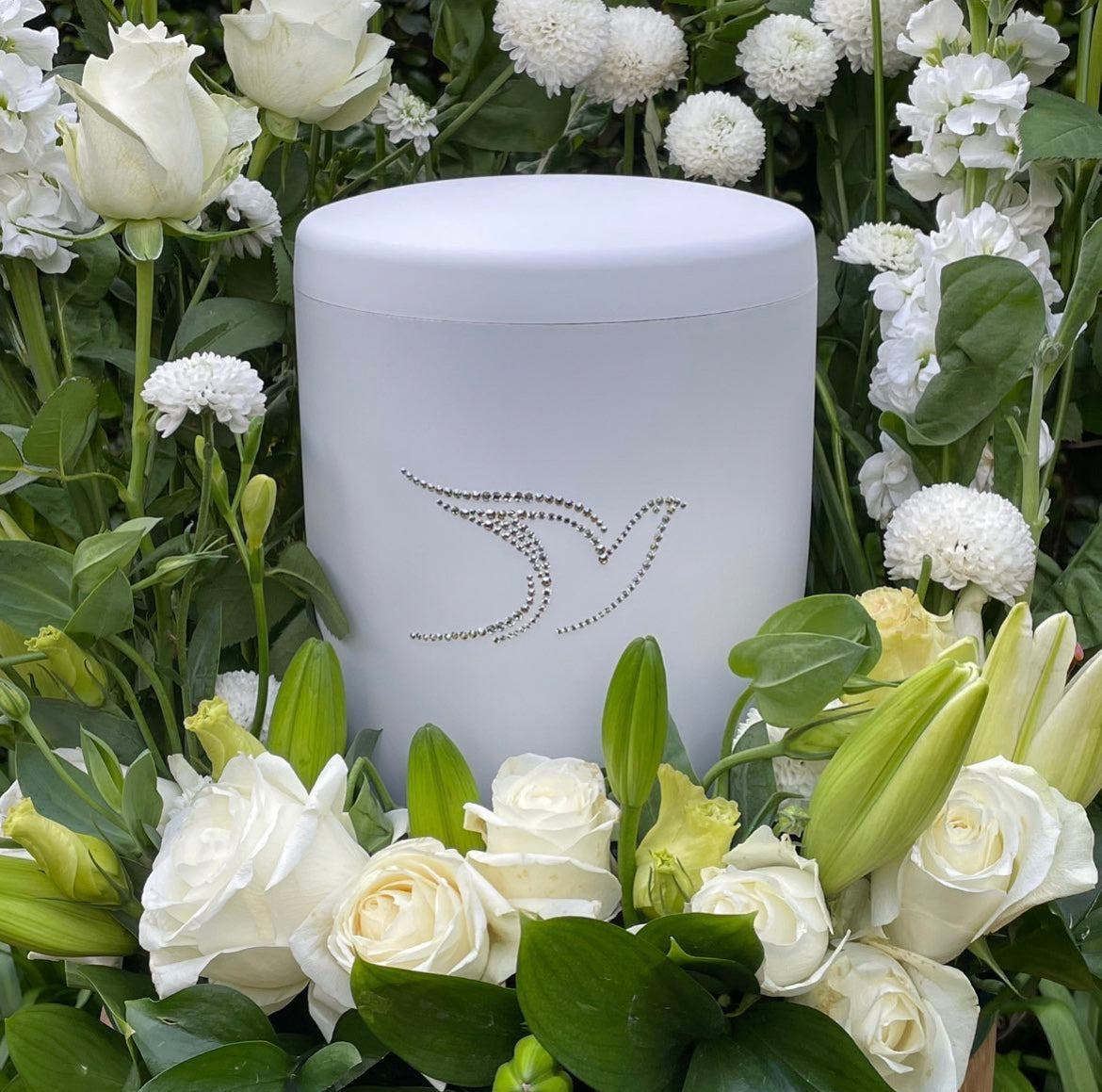 Elegant white urn for ashes adorned with a beautiful dove set in sparkling Swarovski crystals in a bouquet of white lilies and roses.