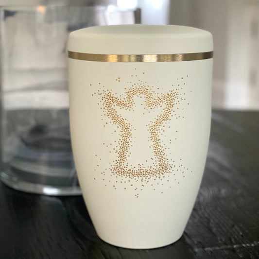 Beautiful ivory coloured urn for ashes with the outline of an angel hand-painted with golden dots on a black table.