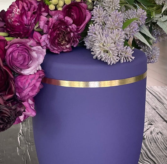 Beautiful violet coloured funeral urn with with a brushed gold band and a colourful flower bouquet.