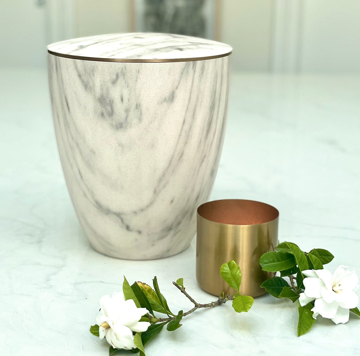 Stunning white marble imitation urn for ashes with a delicate gold band on a marble bench with golden candle and flowers.
