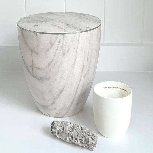 Stunning white marble imitation urn for ashes with a delicate silver band on white shelf next to white devotional candle.