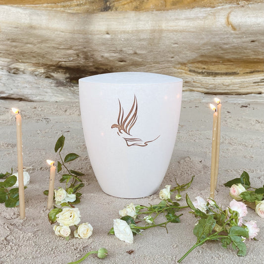 This beautiful off-white urn for human ashes is engraved with an angel in a soft natural colour. The urn is sitting on the beach surrounded by white flowers and devotional candles.