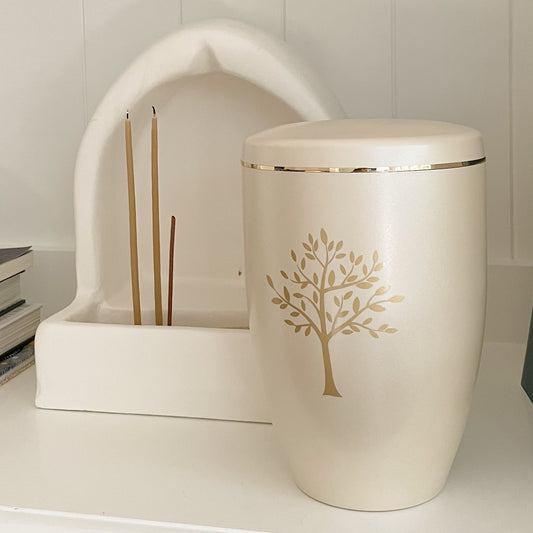 Off-white slightly pearlised cremation urn with an elegant tree of life. This beautiful urn is sitting next to a small white altar with devotional candles.
