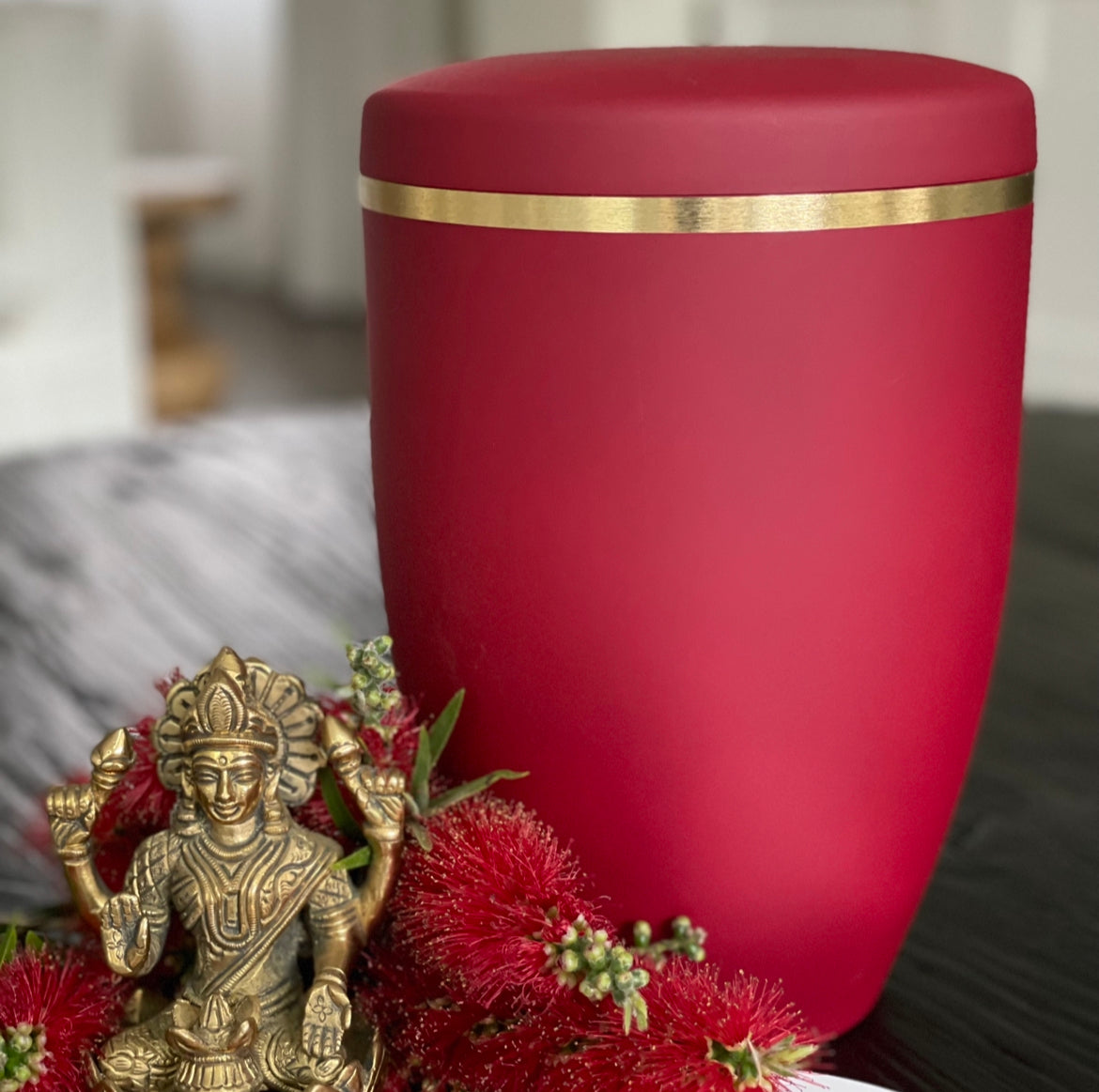 Beautiful ruby coloured urn for ashes with a delicate brushed gold band sat next to a golden Indian statue and Australian bottlebrush branches.