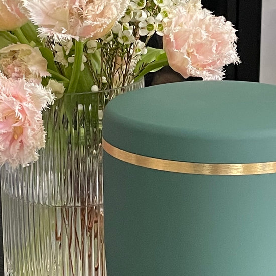 Beautiful sage coloured urn for ashes with a delicate brushed gold band next to beautiful light pink and green flowers.