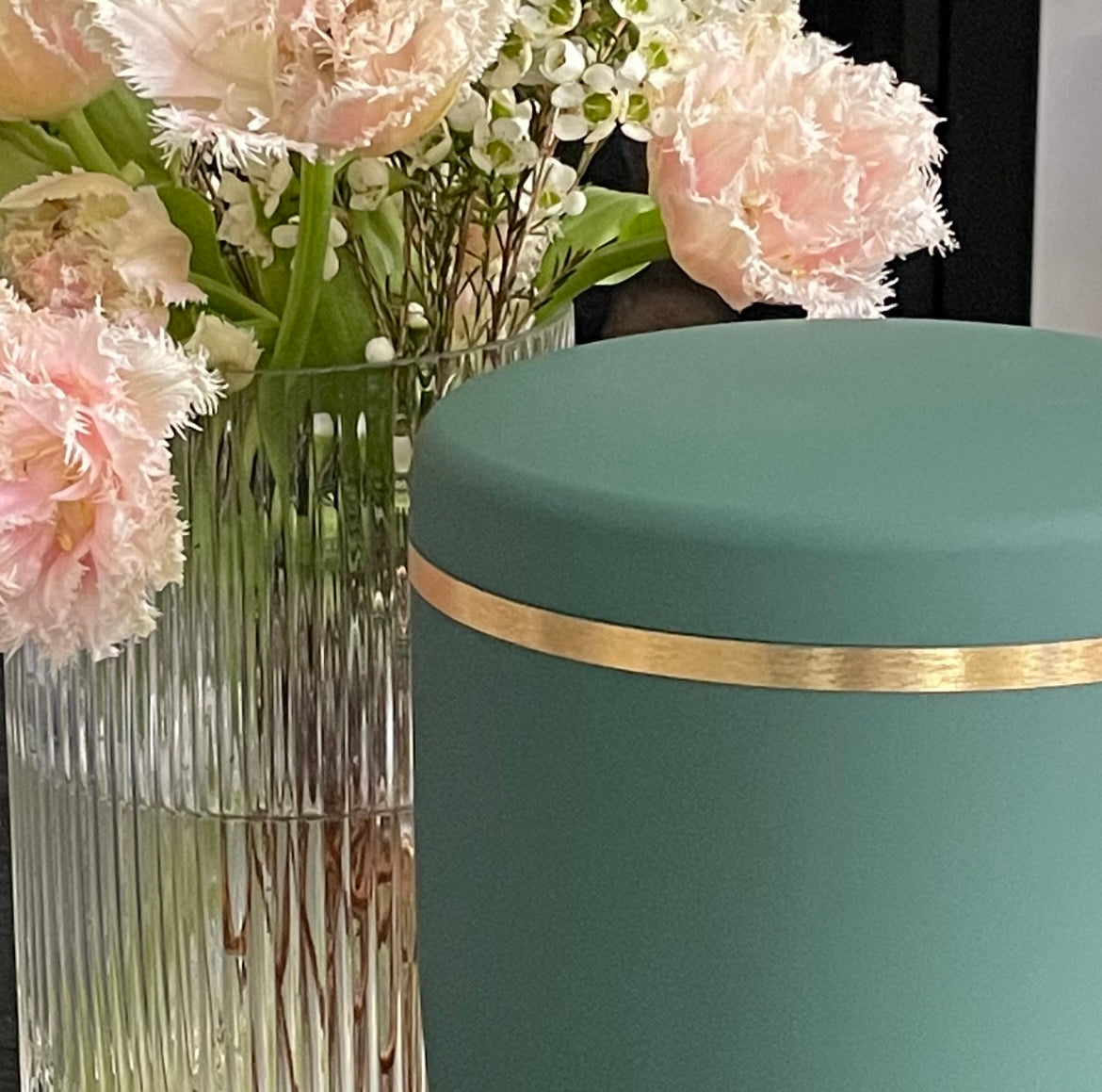 Beautiful sage coloured urn for ashes with a delicate brushed gold band next to beautiful light pink and green flowers.