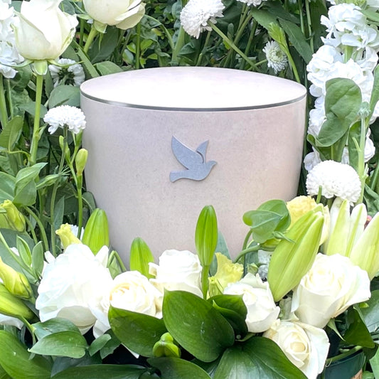 Stunning off-white coloured urn for ashes with a symbol of a silver dove set in a beautiful bouquet of white lilies and roses.