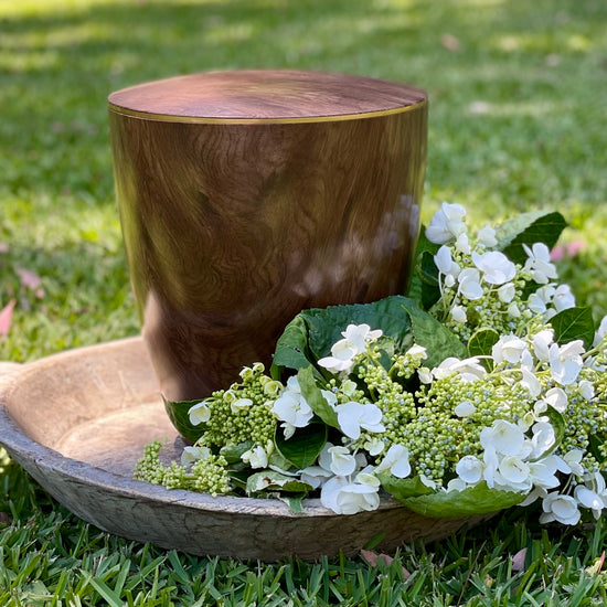 Stunning walnut wood imitation urn with a delicate gold coloured ring sitting on a lawn with an arrangement of white flowers. 