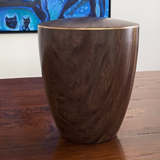 Stunning walnut wood imitation urn with a delicate gold ring sitting on an antique table with a blue painting in the background.