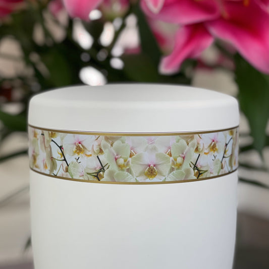 Close-up of a white urn for ashes featuring a beautiful photographic band of white orchids. A beautiful bunch of pink orchids in the background.
