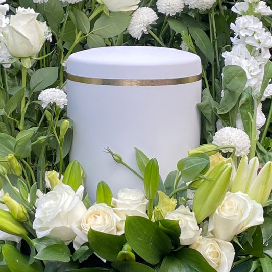 Beautiful white urn for ashes with an elegant brushed gold band set in a beautiful flower wreath of white flowers and foliage.