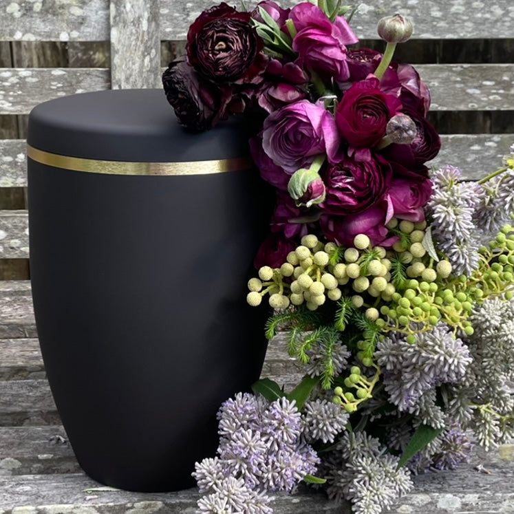 Simple and dignified black urn for ashes with a brushed gold band. The cremation urn sits on a wooden bench with a beautiful flower arrangement of pink and purple flowers and green foliage.