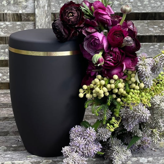Simple and dignified black urn for ashes with a brushed gold band.  The cremation urn sits on a wooden bench with a beautiful flower arrangement of pink and purple flowers and green foliage.