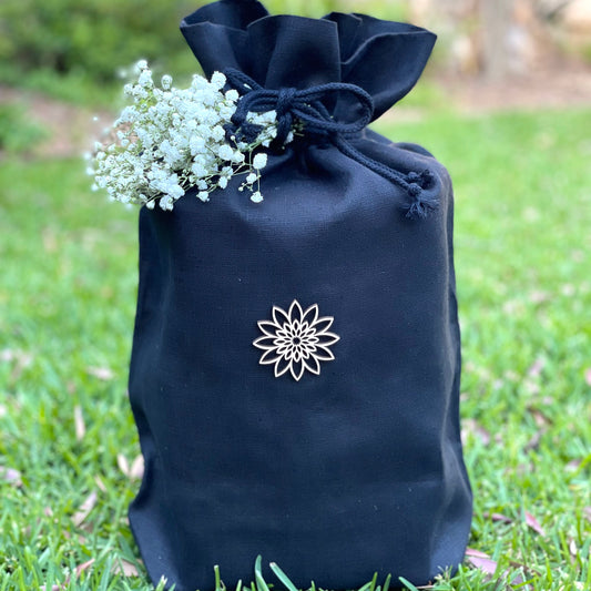 Simple and beautiful black fabric urn for ashes made of the finest ecological Italian linen and adorned with a delicate wooden ornament and delicate white flowers tied to its bow sitting in a garden..