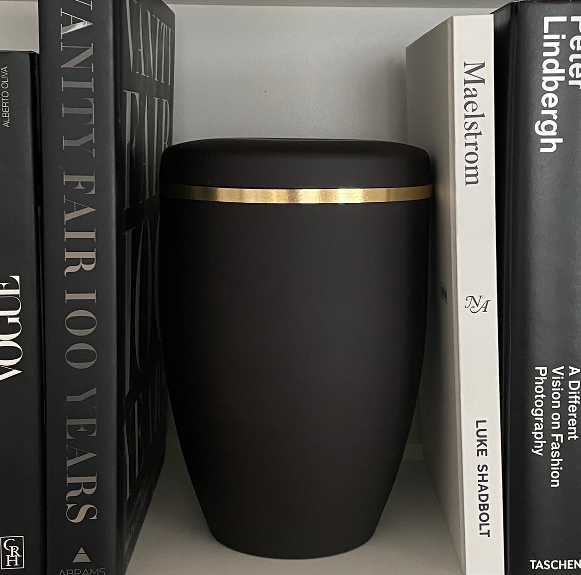 Simple and dignified black urn for ashes with a brushed gold band. The black urn sits on a bookshelf next to black and white books.