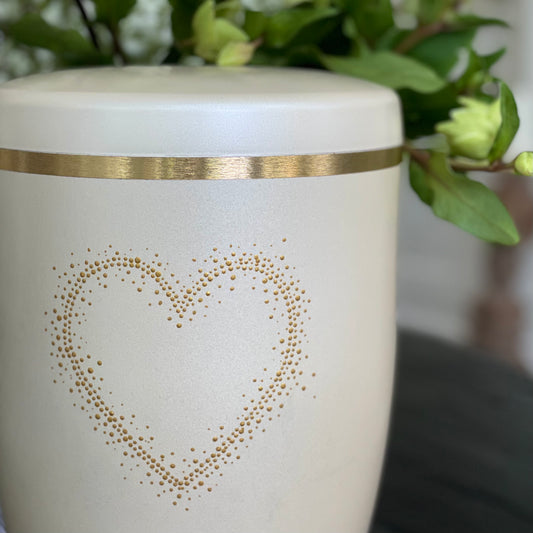 Beautiful hand-painted cream-coloured urn for ashes with a soft pearlised sheen. The urn, which sits on a black table with white flowers in the background, is adorned with the silhouette of a golden heart hand-painted with delicate dots. 