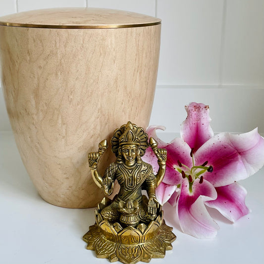 Beautiful realistic looking wood imitation urn for ashes in s blonde maple colour sitting next to a bronze Hindu Goddess and a pink orchid.