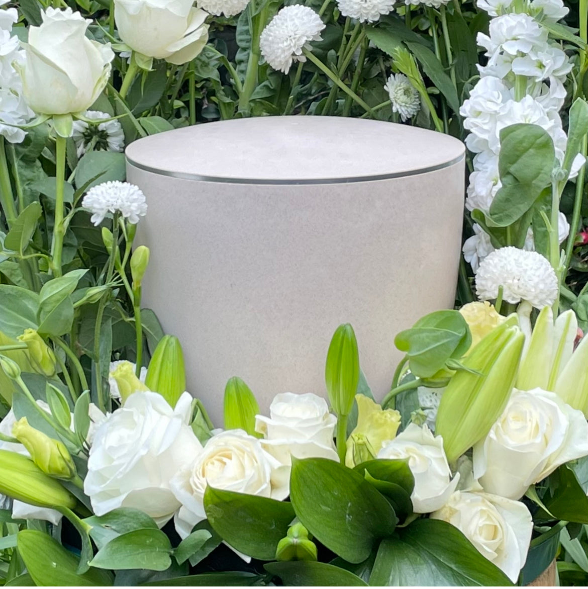 Stunning off-white coloured cremation urn with a delicate silver ring set in a beautiful flower arrangement of white flowers and foliage..