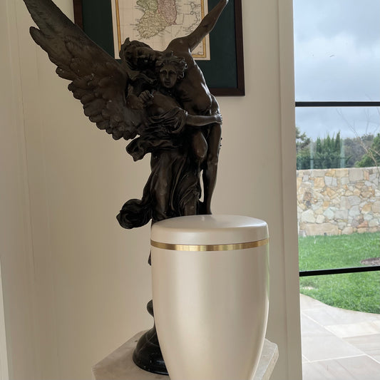Stunning cream coloured funeral urn with an angel statue in the background. an elegant slightly pearlised sheen and a brushed golden band. The urn sits on a marble pillar