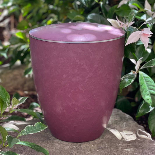 Beautiful soft rose coloured funeral urn with a delicate silver band sitting on a stone with garden flowers.