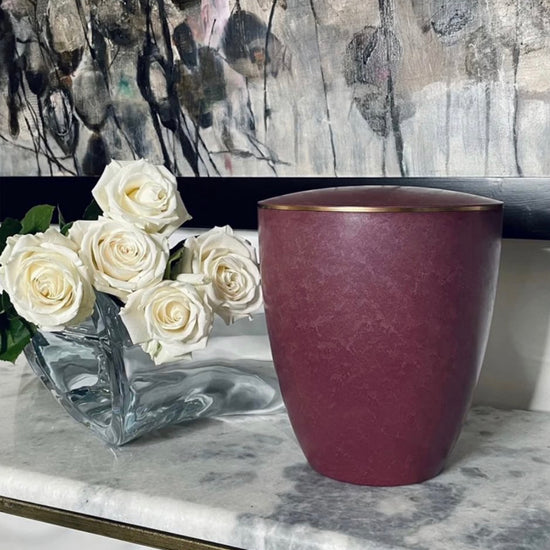 Beautiful urn for ashes in a vibrant rose colour embellished with a delicate 3mm gold ring siting on a marble console.