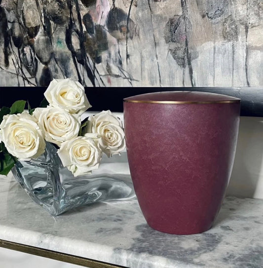 Beautiful urn for ashes in a vibrant rose colour embellished with a delicate 3mm gold ring siting on a marble console.