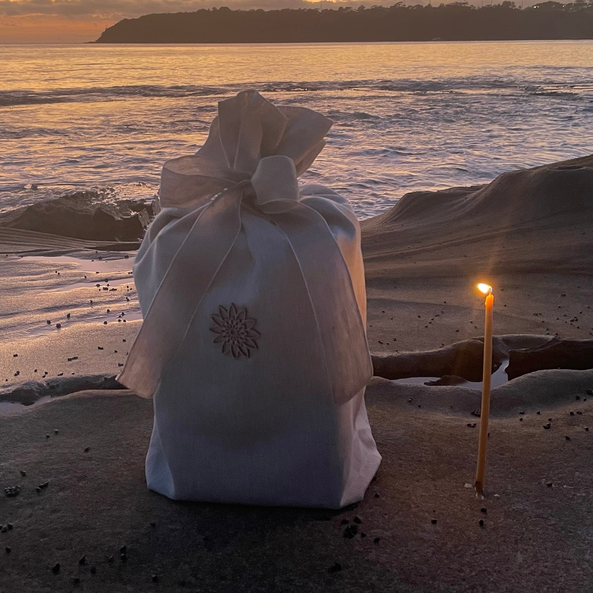 White linen funeral urn sitting on a rock at sunrise with the ocean in the background a burning devotional candle.