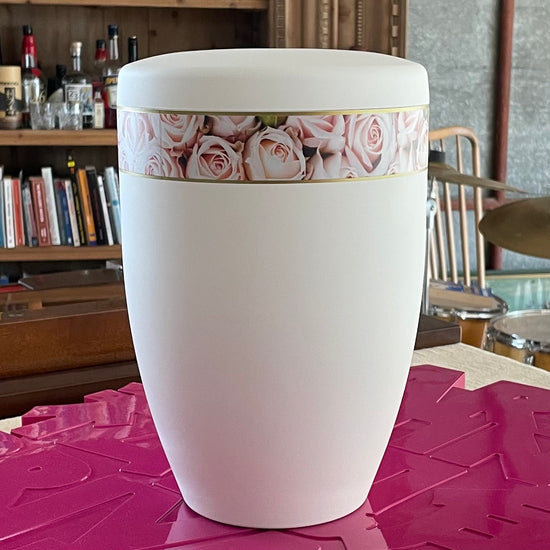 Beautiful white urn for ashes with its band of delicate pink roses sitting on pink artwork.