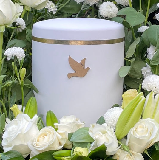 Beautiful white urn for ashes with a golden dove and a delicate brushed gold ring. set in an elegant flower arrangement of white flowers and foliage.