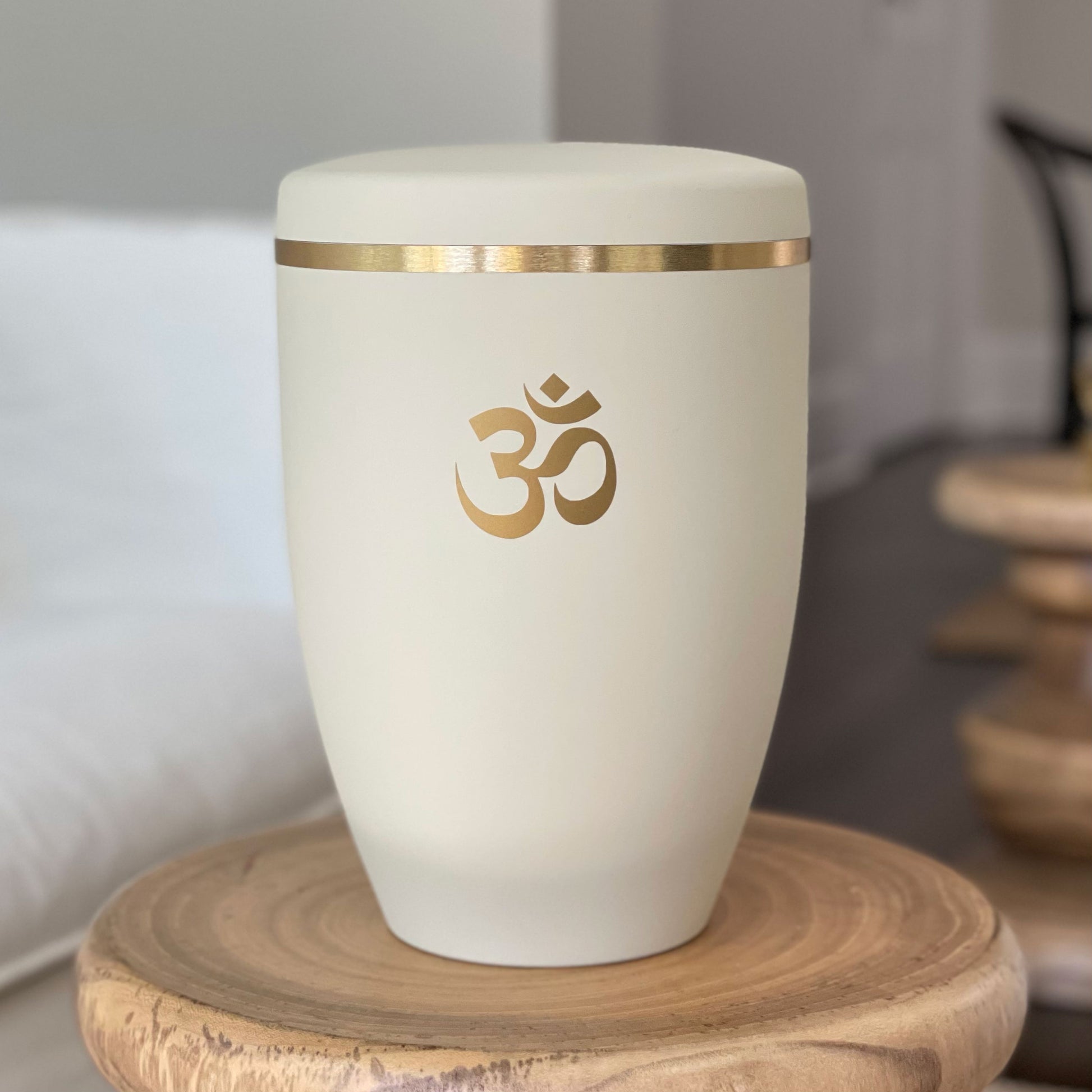 Beautiful ivory coloured funeral urn with a brushed gold band and golden om symbol sitting on a small wooden table with white furniture in the background.