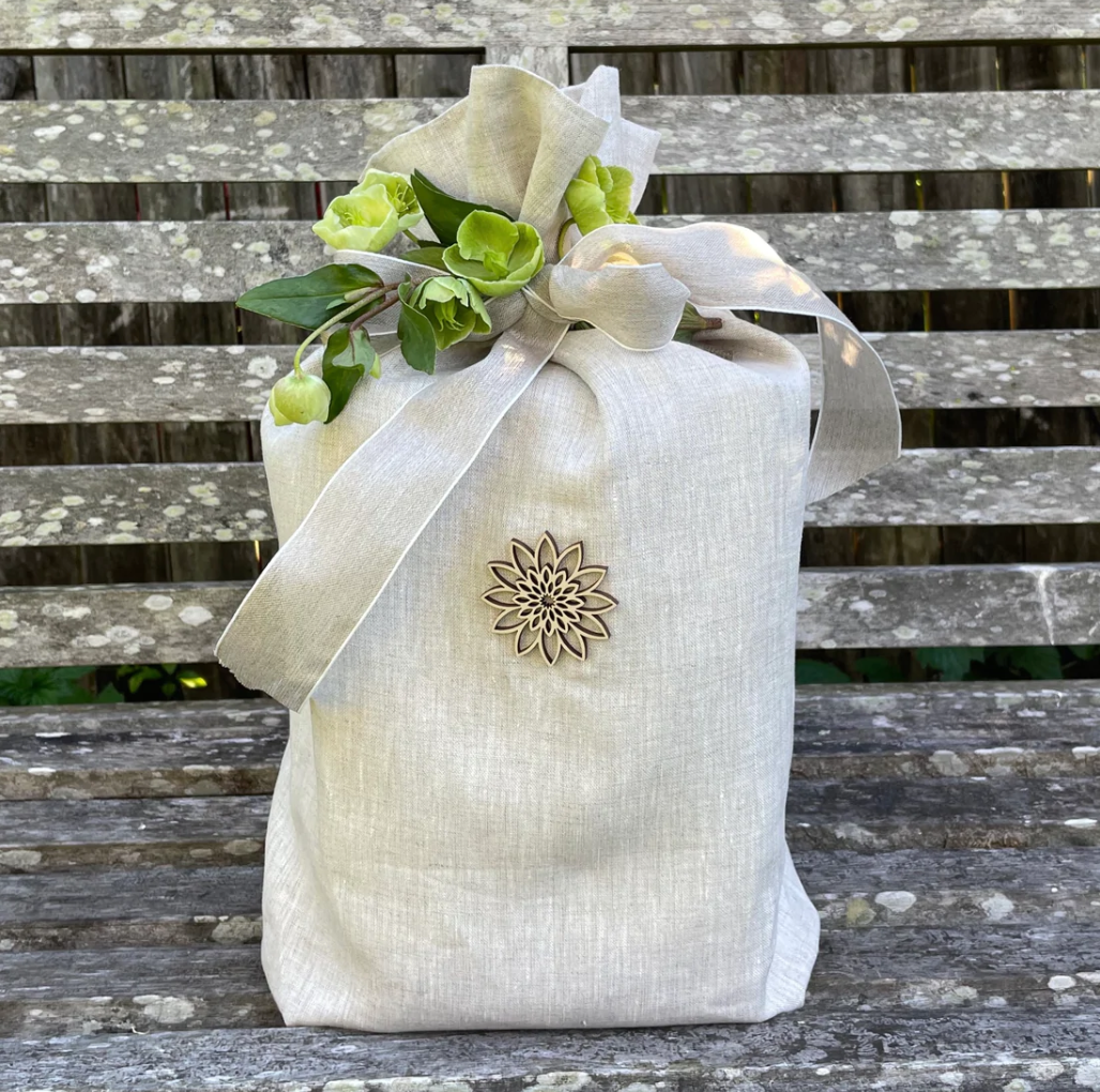 Beautiful oatmeal coloured linen funeral urn with wooden ornament and delicate garden blooms tied to it with a linen bow. The urn is sitting on a weathered wooden bench.