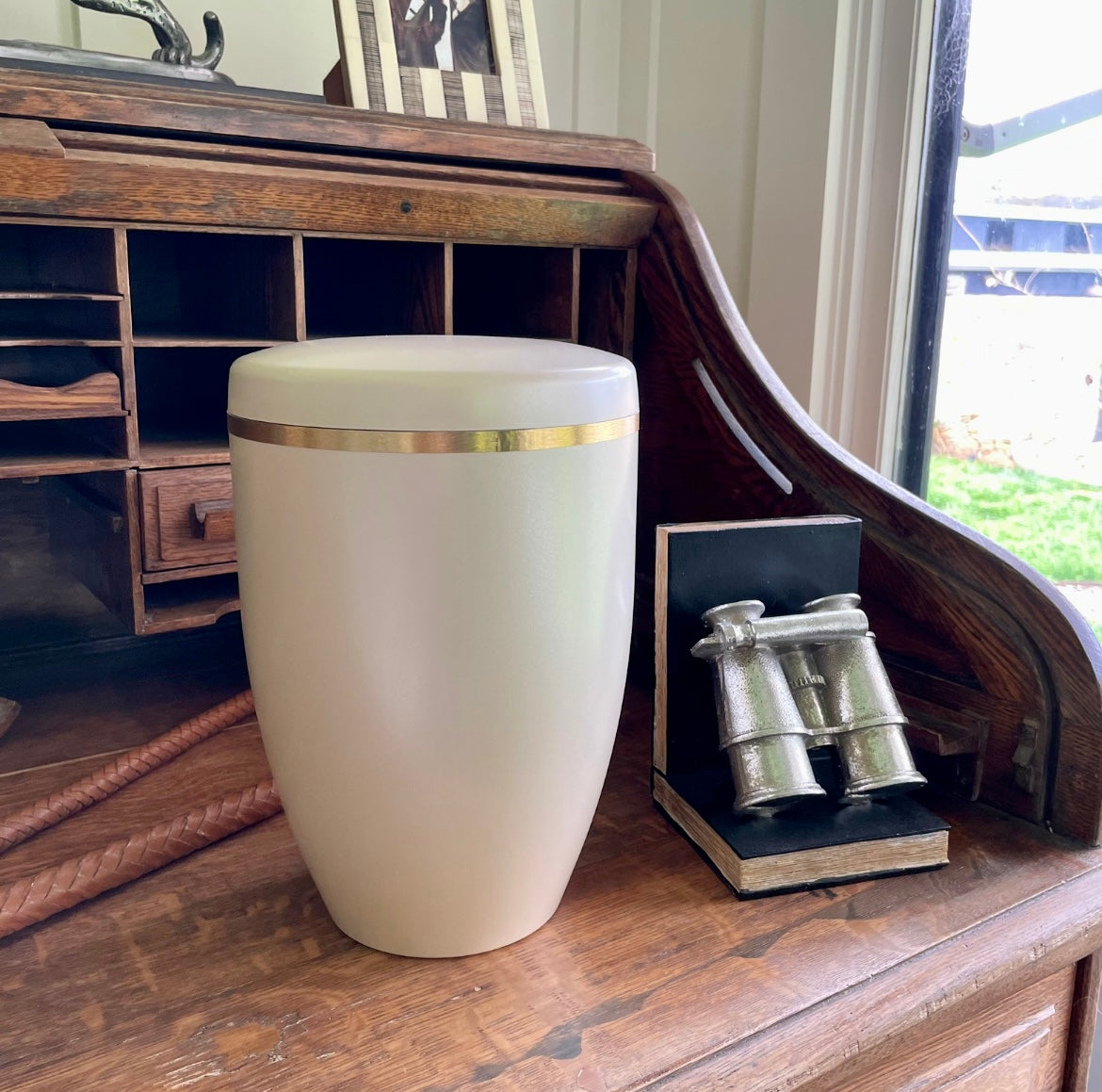 Beautiful cream coloured urn for ashes with a slightly pearlised sheen and a brushed golden band. The urn sits on an antique wooden desk.